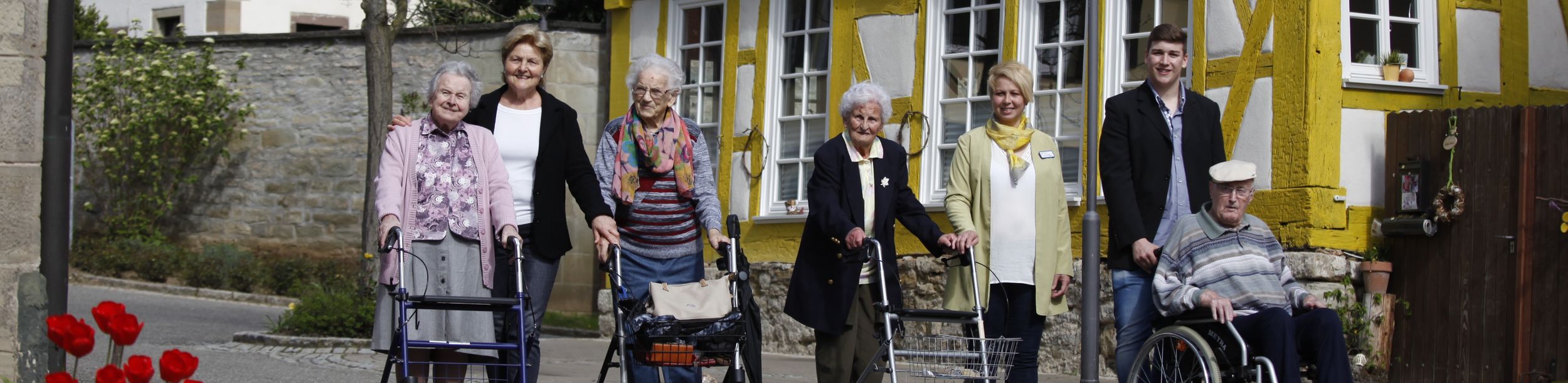 Seniorinnen und Senioren mit Rollatoren und Rollstuhl neben Pflegenden vor Fachwerkhaus