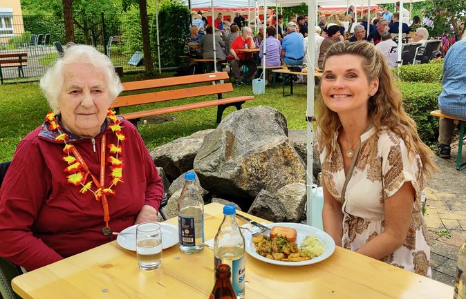 Ältere Frau mit Hawaii-Blumenkranz und lächelnde Begleiterin am Festtisch